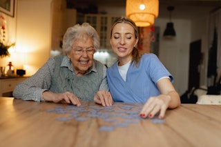 Aînée atteinte d’Alzheimer faisant un casse-tête à domicile avec une soignante, dans le cadre de soins adaptés.