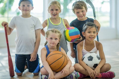 A multi-ethnic group of elementary school students are in indoors in a fitness center. They are wearing athletic clothing. They are sitting on the floor holding various sports equipment, including a basketball, soccer ball, volleyball, hockey stick, and baseball bat.